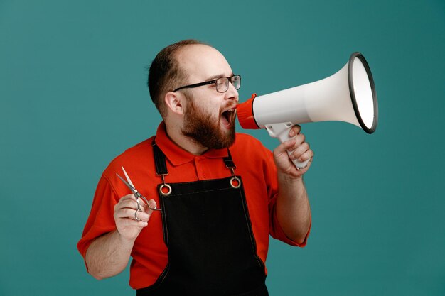 Jeune barbier sérieux portant des lunettes chemise rouge et tablier de coiffeur tenant des ciseaux regardant le côté parlant dans le haut-parleur isolé sur fond bleu