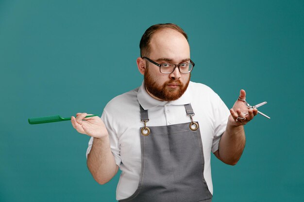 Jeune barbier sérieux en uniforme et lunettes regardant la caméra tenant un peigne teaser et des ciseaux isolés sur fond bleu