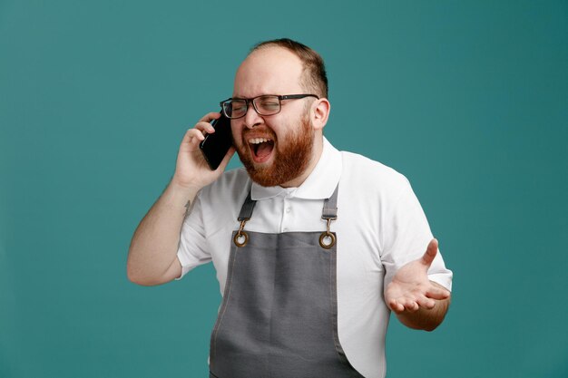 Jeune barbier stressé en uniforme et lunettes parlant au téléphone criant les yeux fermés montrant la main vide isolée sur fond bleu