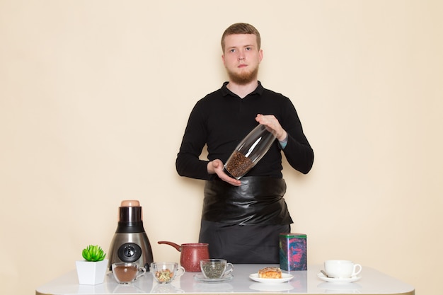 Jeune barista en costume de travail noir avec des ingrédients et du matériel de café graines de café brun sur blanc