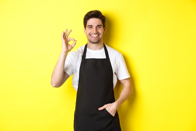 Jeune barista souriant en tablier noir montrant un signe d'accord, recommandant un café ou un restaurant, debout sur fond jaune