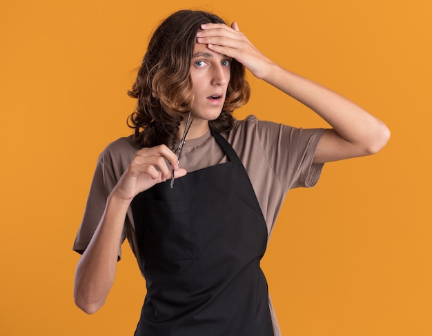 Jeune beau barbier concerné portant des ciseaux tenant des uniformes mettant la main sur le front