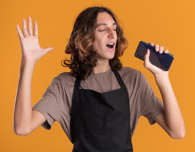Jeune et beau barbier joyeux en uniforme gardant la main dans l'air tenant un téléphone portable l'utilisant comme microphone chantant les yeux fermés isolés sur un mur orange