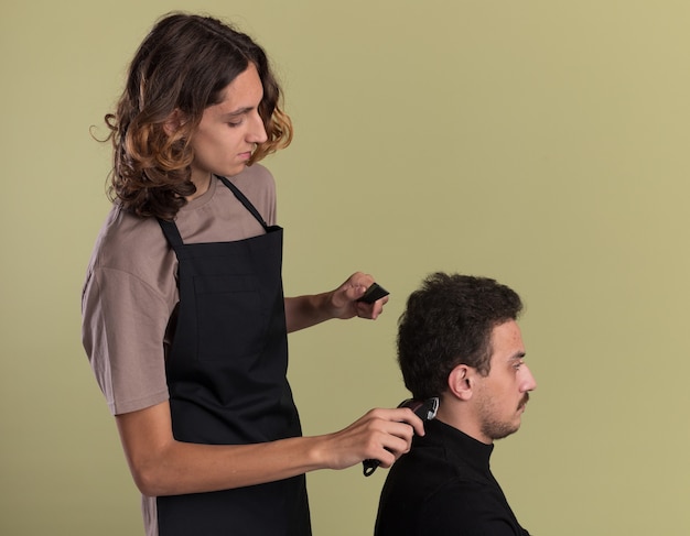 Jeune Beau Barbier En Uniforme Debout En Vue De Profil Faisant Une Coupe De Cheveux Pour Son Jeune Client Photo gratuit