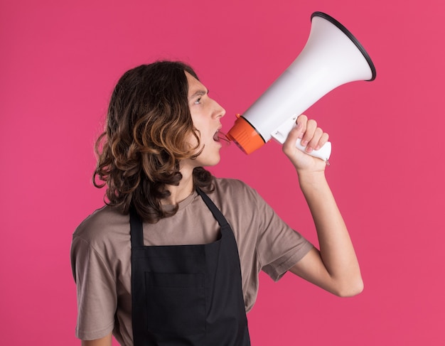 Photo gratuite jeune beau barbier en uniforme tournant la tête à l'autre en levant parler par le haut-parleur isolé sur le mur rose