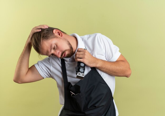 Jeune beau coiffeur tenant une tondeuse à cheveux mettant la main sur la tête avec les yeux fermés isolé sur fond vert olive