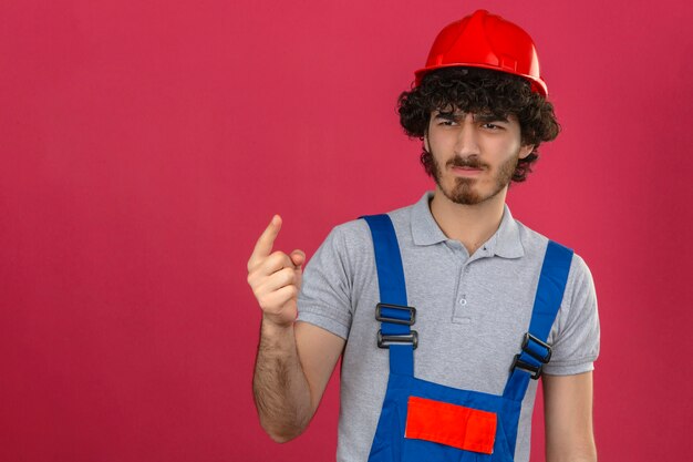 Jeune beau constructeur barbu portant des uniformes de construction et un casque de sécurité pointant avec le doigt vers le haut et l'expression en colère ne montrant aucun geste sur un mur rose isolé