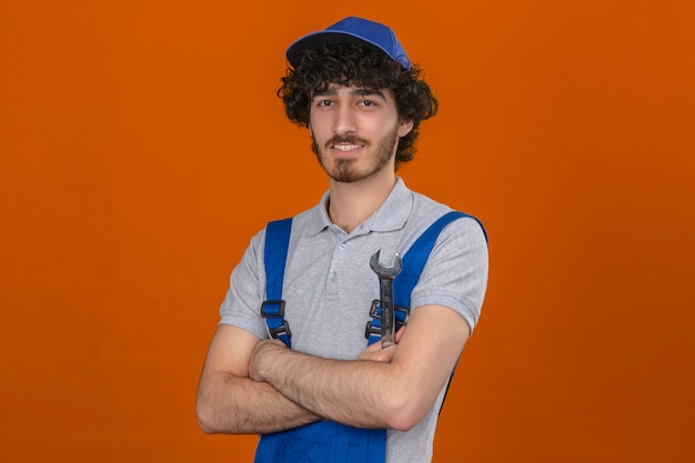 Photo gratuite jeune beau constructeur barbu portant des uniformes de construction et une casquette debout avec les bras croisés souriant sur mur orange isolé