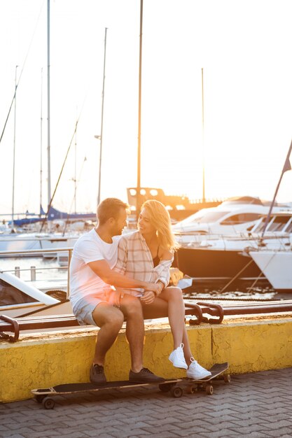 Photo gratuite jeune beau couple assis au bord de mer, souriant, planche à roulettes.