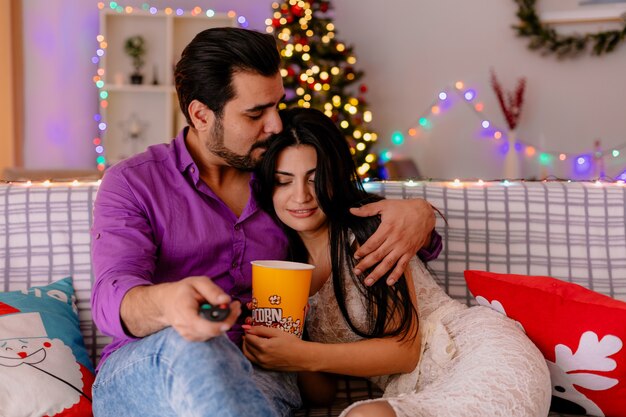 Jeune et beau couple assis sur un canapé homme et femme avec seau de pop-corn à regarder la télévision ensemble heureux en amour dans une chambre décorée avec arbre de Noël en arrière-plan