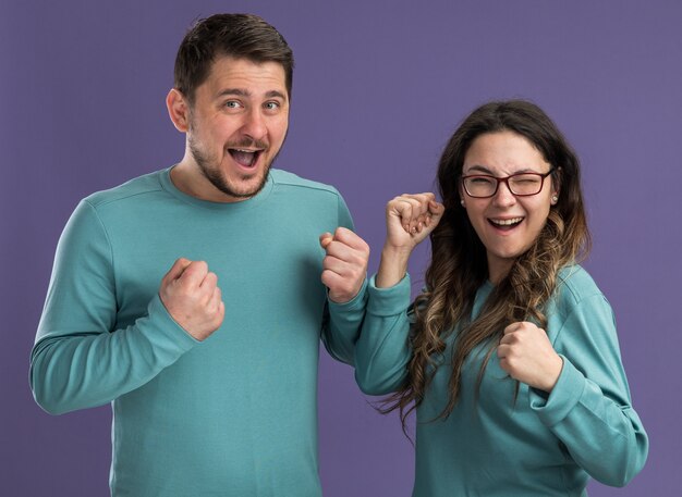 Jeune beau couple en bleu vêtements décontractés homme et femme heureux et excités serrant les poings heureux en amour debout sur le mur violet