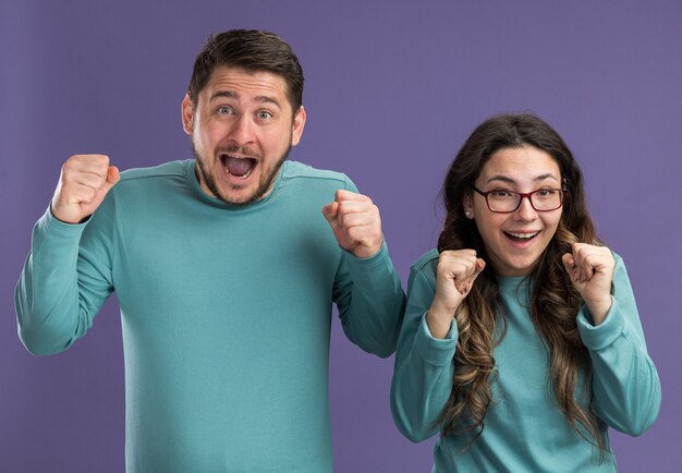 Jeune beau couple en bleu vêtements décontractés homme et femme serrant les poings fou heureux et excité debout sur le mur violet