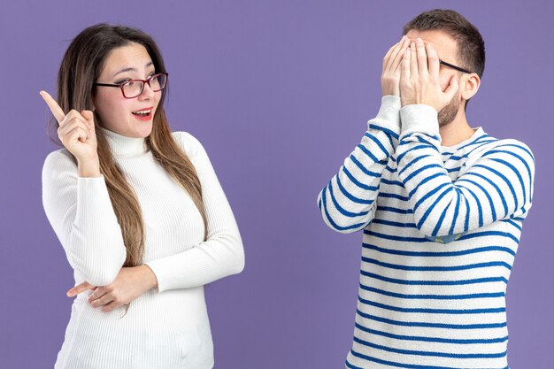 Jeune beau couple dans des vêtements décontractés femme heureuse et surprise en regardant son petit ami qui couvrant le visage avec des palmiers Saint Valentin debout sur un mur violet