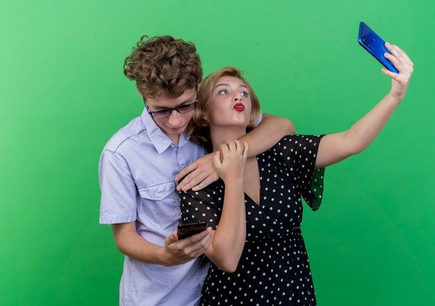 Jeune beau couple debout ensemble à l'aide de téléphone mobile prenant selfie soufflant baiser sur mur vert