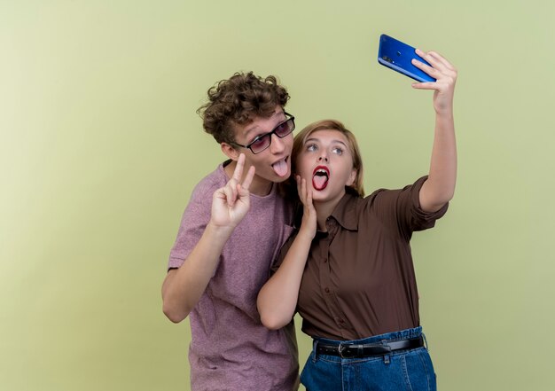 Jeune beau couple ensemble à l'aide de téléphone mobile prenant selfie smiling sticking out tongue et montrant v-sign sur la lumière