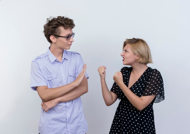 Jeune beau couple femme en colère regardant avec les poings fermés à son petit ami confus qui montrant le geste d'arrêt sur un mur blanc