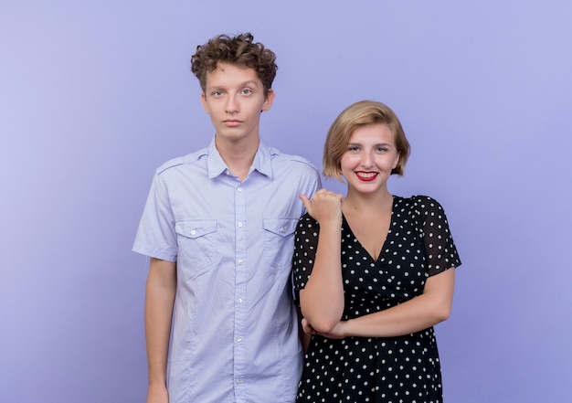 Photo gratuite jeune beau couple femme heureuse pointant sur son petit ami qui avec un visage sérieux debout sur un mur bleu