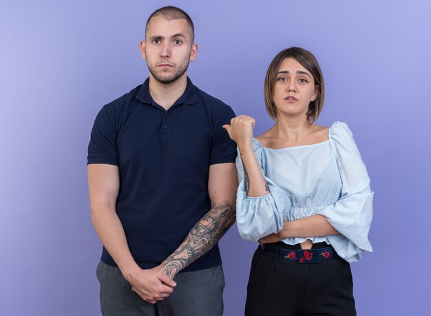 Jeune beau couple femme mécontente montrant son petit ami confus debout sur le mur bleu