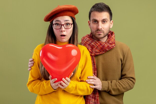 Jeune beau couple femme surprise en béret avec ballon en forme de coeur et homme avec écharpe surpris célébrant la Saint-Valentin debout sur le mur vert