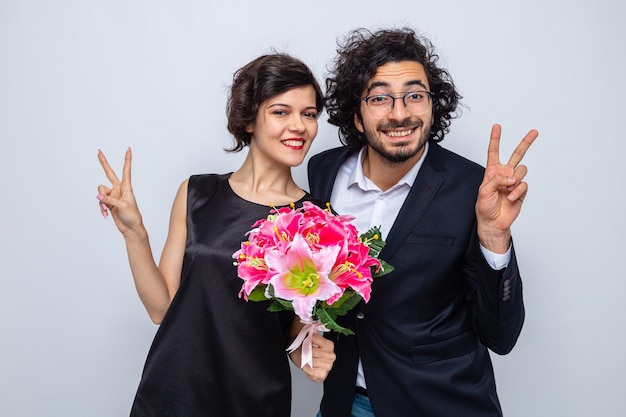 Jeune beau couple heureux homme et femme avec bouquet de fleurs regardant la caméra en souriant joyeusement montrant les pouces vers le haut célébrant la journée internationale de la femme le 8 mars debout sur fond blanc