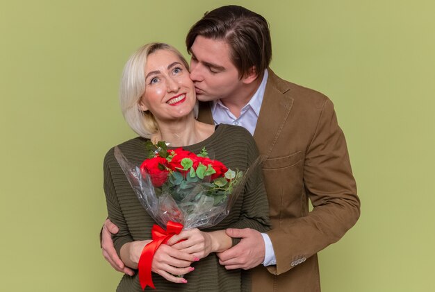 Jeune beau couple heureux homme et femme avec bouquet de roses rouges embrassant heureux en amour ensemble célébrant la journée internationale de la femme debout sur le mur vert