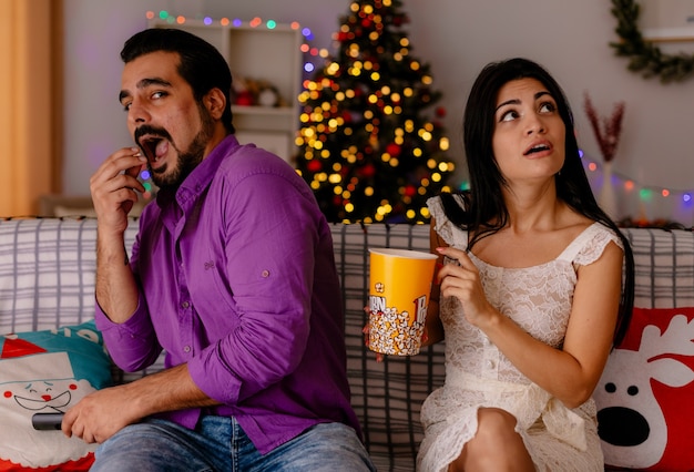 Jeune et beau couple heureux homme gai et femme perplexe avec un seau de pop-corn regardant la télévision ensemble dans une pièce décorée avec un arbre de Noël dans le mur