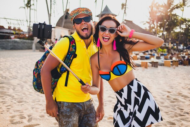 Jeune beau couple hipster amoureux sur la plage tropicale, prenant selfie photo sur smartphone, lunettes de soleil, tenue élégante, vacances d'été, s'amuser, souriant, heureux, coloré, émotion positive