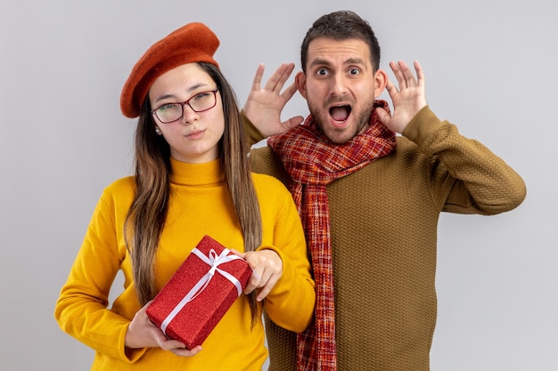 Jeune beau couple homme excité et femme souriante en béret tenant présent avec sourire sur le visage célébrant la Saint-Valentin debout sur un mur blanc