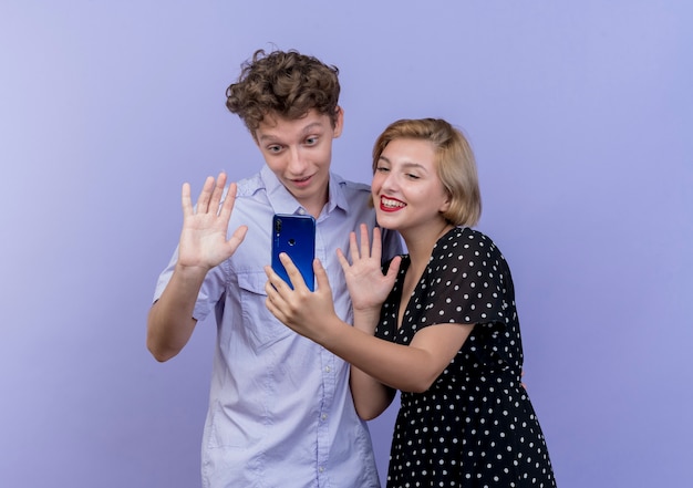 Jeune beau couple homme et femme ayant un appel vidéo en agitant les mains et souriant sur bleu