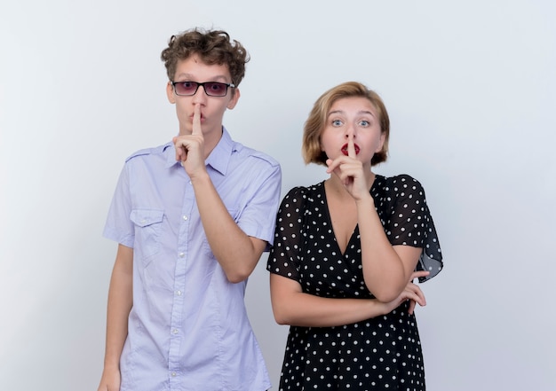 Jeune beau couple homme et femme debout à côté de l'autre faisant un geste de silence avec les doigts sur les lèvres sur un mur blanc