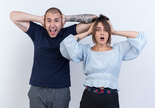 Jeune beau couple homme et femme étonné et surpris avec les mains sur la tête debout sur un mur blanc