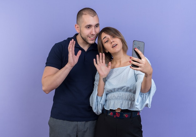 Jeune beau couple homme et femme heureux et positif souriant joyeusement faisant selfie à l'aide de smartphones agitant les mains debout sur le mur bleu