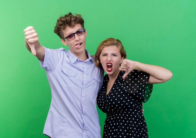 Jeune beau couple homme et femme mécontent montrant les pouces vers le bas debout sur le mur vert