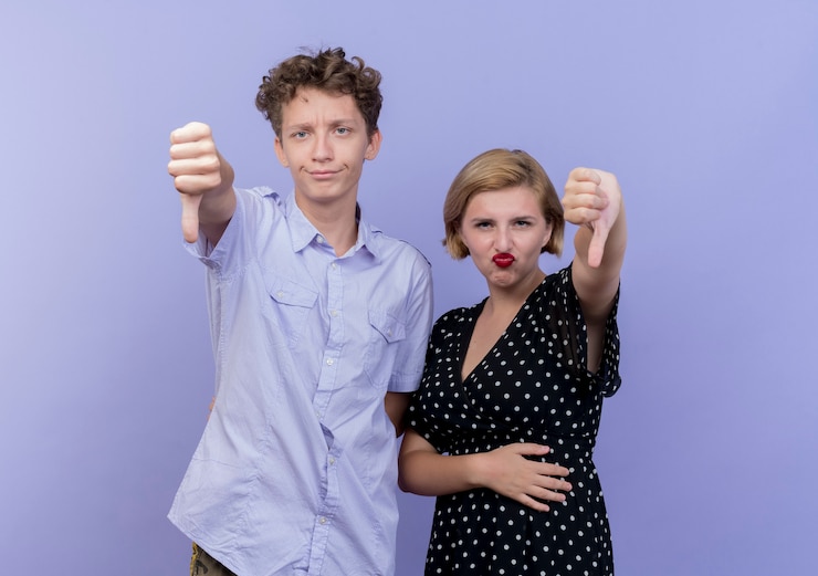 Photo gratuit jeune beau couple homme et femme à la mécontentement montrant les pouces vers le bas sur le mur bleu