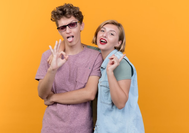 Jeune beau couple homme et femme portant des vêtements décontractés posant et souriant montrant signe ok debout sur un mur orange