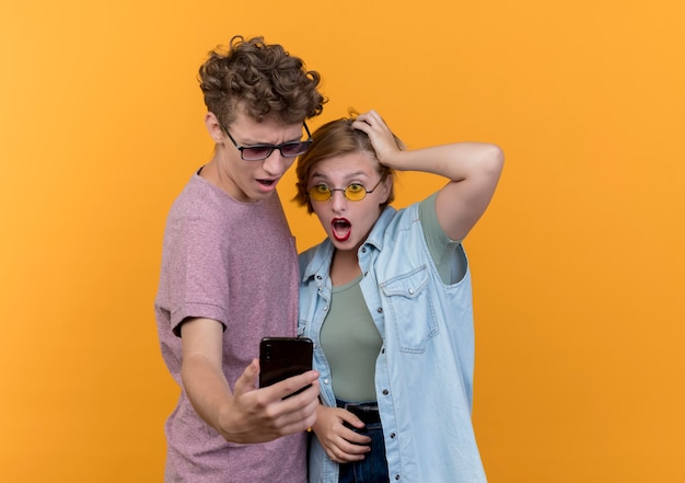 Jeune beau couple homme et femme portant des vêtements décontractés regardant smartphone surpris et confus debout sur le mur orange
