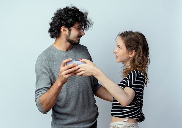 Jeune beau couple homme et femme regardant les uns les autres tenant un smartphone en prenant une photo d'eux ensemble debout sur un mur blanc