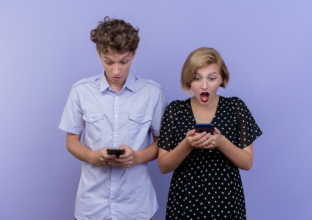 Jeune beau couple homme et femme regardant leurs smartphones surpris debout sur le mur bleu