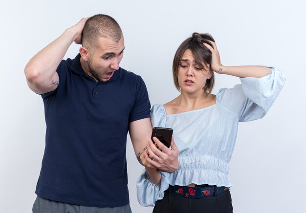 Jeune beau couple homme et femme avec smartphone le regardant confondu avec les mains sur la tête debout