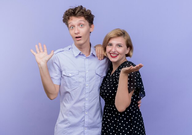 Jeune beau couple homme et femme souriant et écartant les bras sur les côtés debout sur le mur bleu