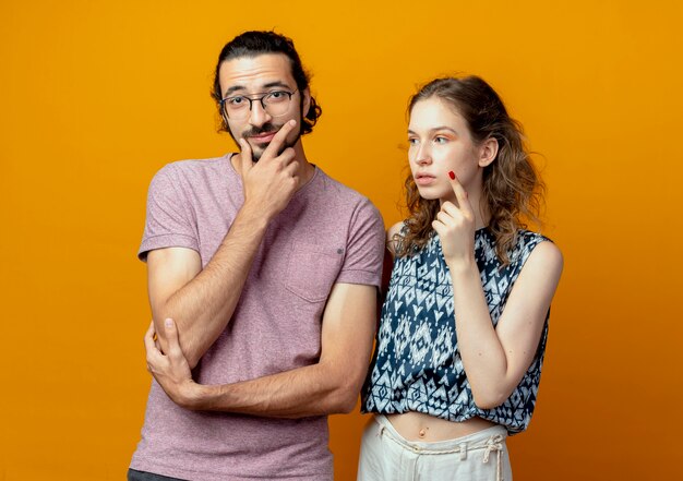 Jeune beau couple homme et femmes à la pensée perplexe debout sur le mur orange