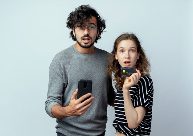 Jeune beau couple homme et femmes regardant la caméra confus man holding smartphone debout à côté de sa petite amie qui détenant une carte de crédit sur fond blanc