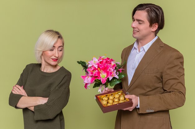 Jeune beau couple homme heureux donnant une boîte de bonbons au chocolat et bouquet de fleurs