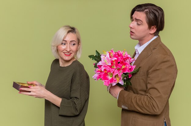 Jeune beau couple homme heureux donnant un bouquet de fleurs à sa petite amie souriante avec boîte de bonbons au chocolat célébrant la journée internationale de la femme debout sur le mur vert
