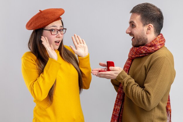 Jeune beau couple homme heureux faisant la proposition avec bague de fiançailles en boîte rouge à sa petite amie confuse en béret pendant la Saint-Valentin debout sur fond blanc