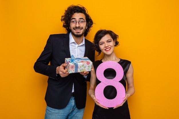 Jeune beau couple homme heureux avec présent et femme avec numéro huit regardant la caméra souriant joyeusement célébrant la journée internationale de la femme le 8 mars debout sur fond orange