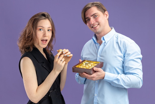 Jeune beau couple homme souriant offrant des bonbons au chocolat à sa charmante petite amie célébrant la Saint-Valentin debout sur le mur bleu