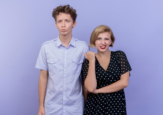 Jeune beau couple mécontent femme pointant sur son petit ami confus debout sur le mur bleu