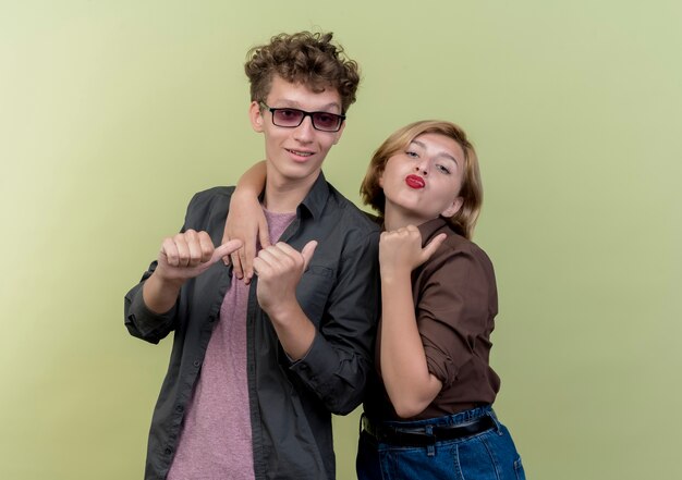 Jeune beau couple portant des vêtements décontractés à la recherche de sourire joyeusement montrant les pouces vers le haut debout sur un mur léger