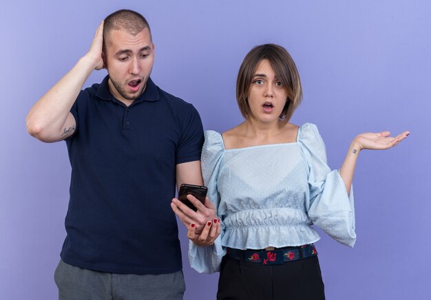 Jeune beau couple en vêtements décontractés homme avec smartphone et femme à la confusion écartant les bras sur les côtés debout
